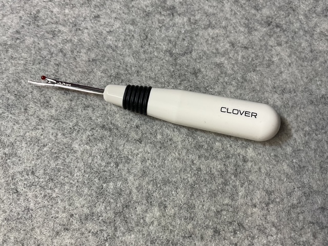 A Clover brand seam ripper with a white handle and a black grip, resting on a gray fabric surface.