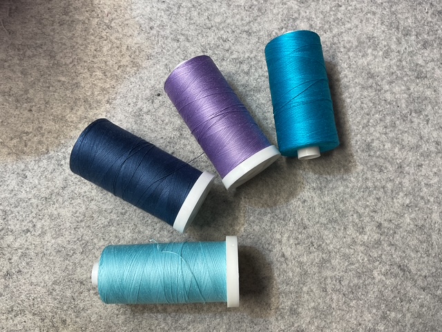 Four spools of thread in shades of dark blue, purple, teal, and light blue, placed on a gray fabric surface.