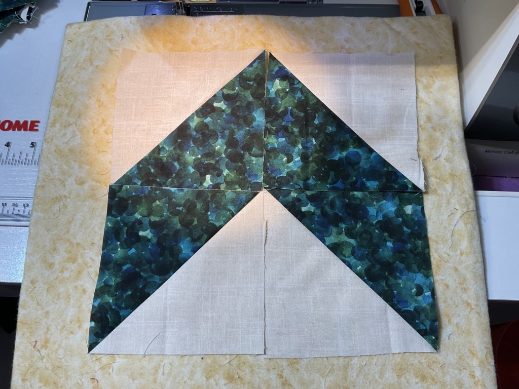 A close-up of a fabric quilt block featuring a geometric design with dark green and blue patterns alongside cream-colored fabric squares. The background is light-colored quilt batting.