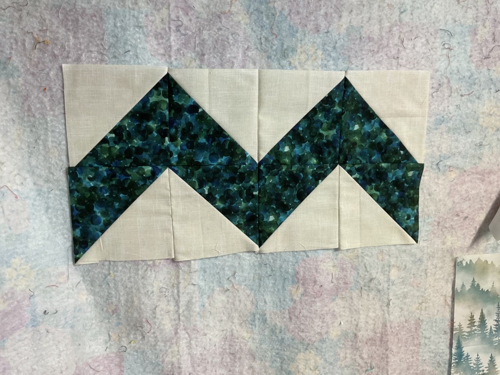 A quilt block featuring a zigzag pattern with dark green and blue prints complemented by light beige fabric.