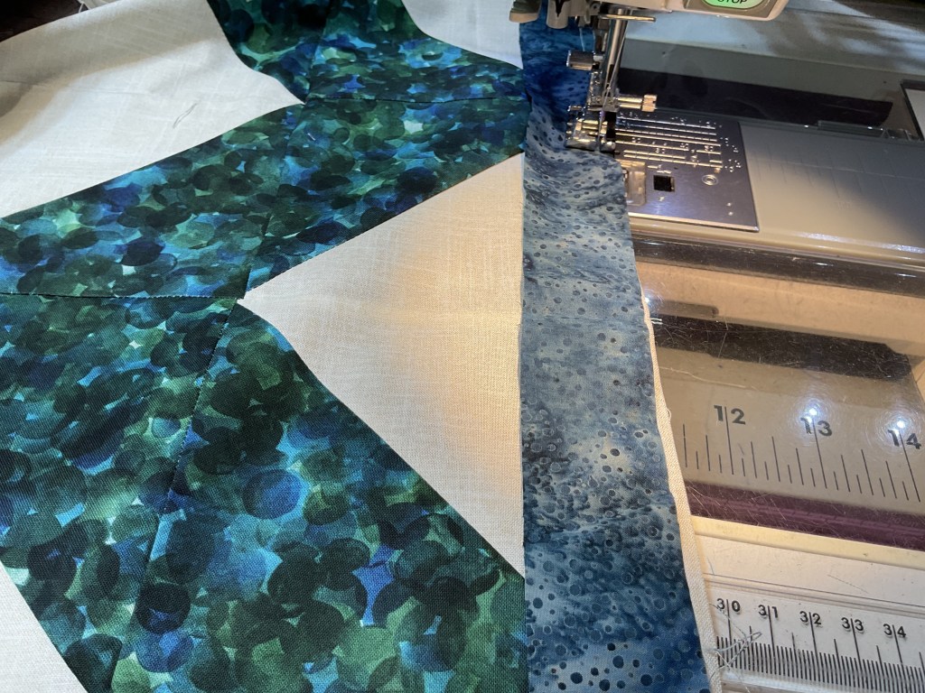 Close-up of a sewing machine working on a quilt with colorful fabric pieces, featuring shades of green and blue, on a light-colored background.