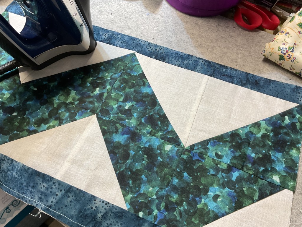 An iron placed on a colorful quilt block featuring green and blue fabric patterns, with unfinished quilt pieces on a sewing surface.