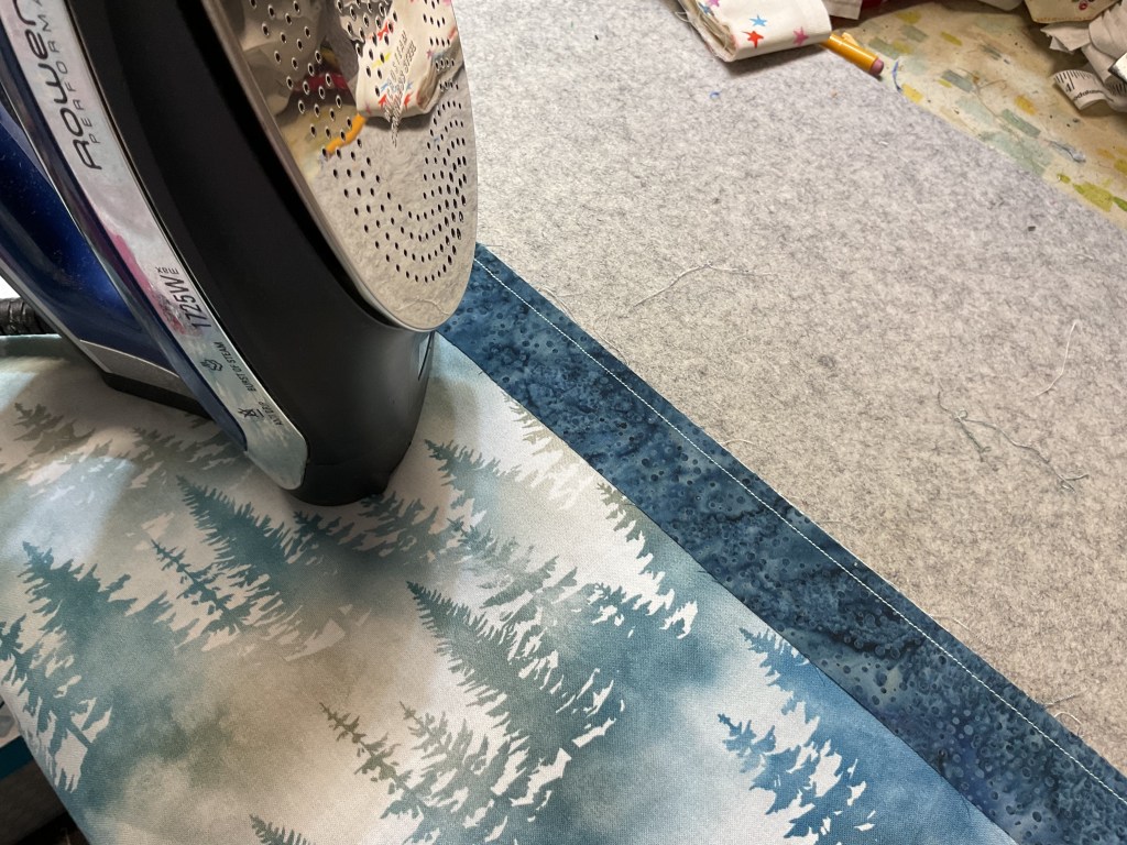 A close-up of an iron positioned on a piece of fabric featuring a forest pattern, with a gray pressing surface underneath.