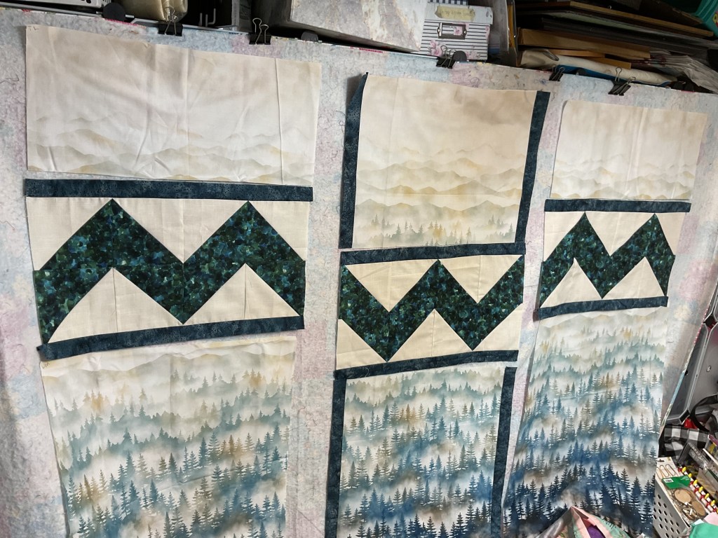 A patchwork quilt in progress featuring layered fabrics with mountainous and forest designs, arranged in a pattern of alternating colors and shapes.