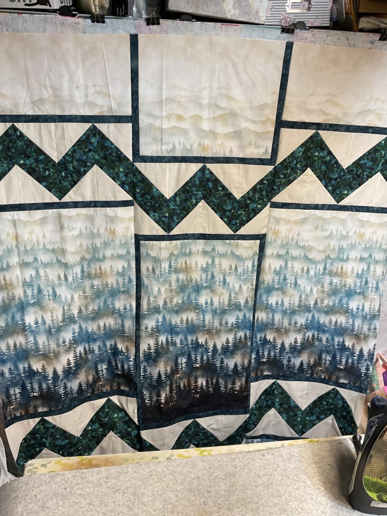 A creatively designed quilt featuring soft, blended colors depicting mountain landscapes and forest silhouettes with a geometric zigzag pattern at the bottom.