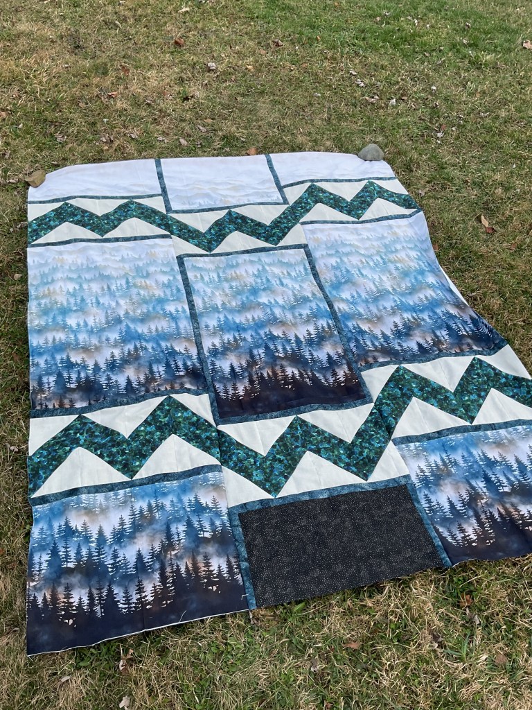 A patchwork quilt laid out on grass, featuring designs of blue and green mountains and forest landscapes.
