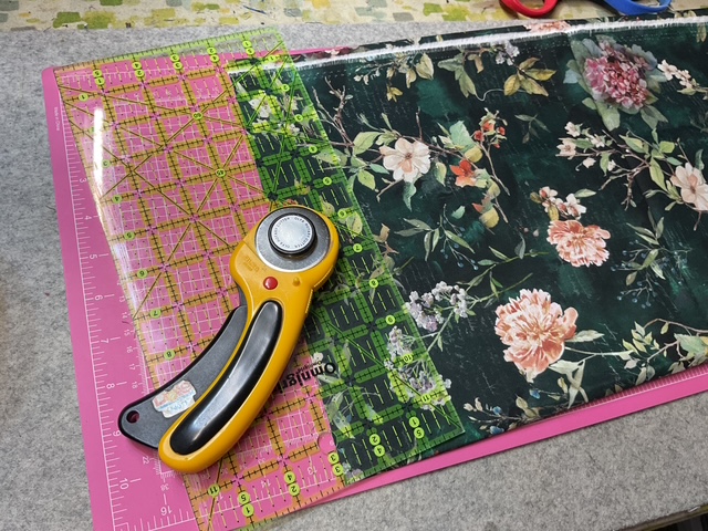 A rotary cutter and a clear acrylic ruler placed on a floral printed fabric, with a pink cutting mat underneath.