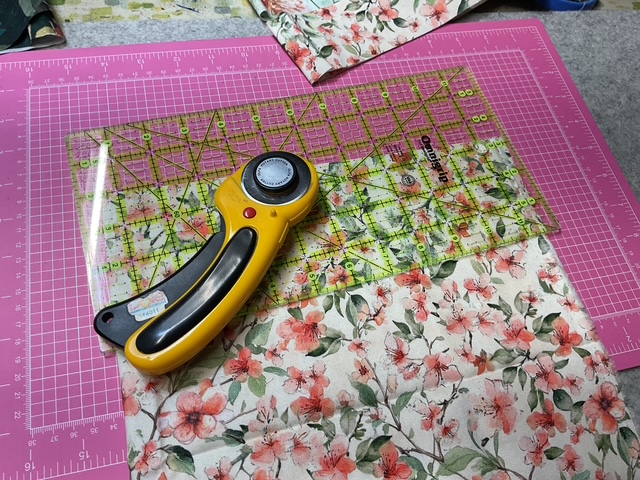 A rotary cutter and a transparent quilting ruler are placed on a floral fabric, set against a pink cutting mat.