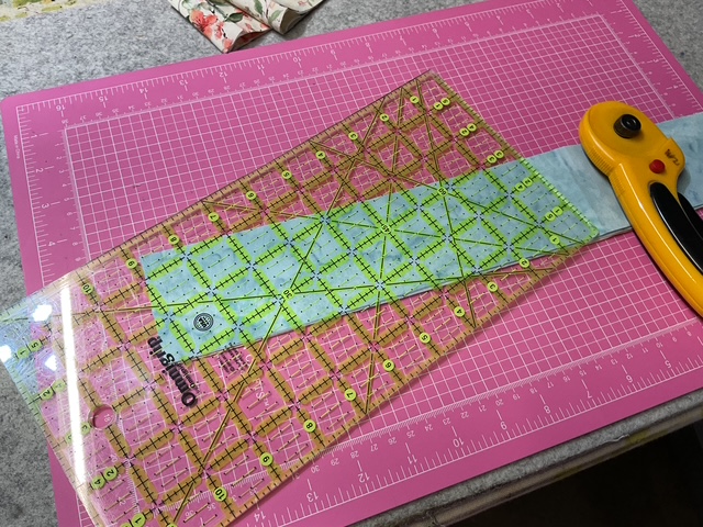 A cutting mat with a transparent quilting ruler placed over a light blue fabric strip and a rotary cutter nearby.