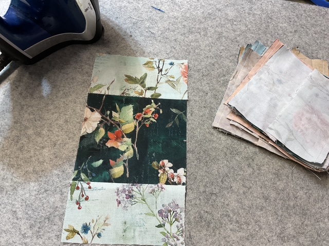 A piece of fabric with floral patterns laid on gray felt, alongside a stack of assorted fabric swatches and an iron.