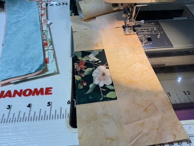 Close-up view of a sewing machine with fabric pieces being stitched together, featuring floral patterns and pastel colors, on a quilting mat.