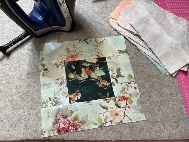 A square fabric patch featuring floral designs laid out on a work surface, with an iron and additional fabric pieces beside it.