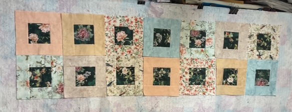 A quilt layout featuring a series of fabric squares arranged in a grid pattern. Each square has a floral print center surrounded by solid pastel colors. The layout includes a mix of peach, beige, and blue fabrics.
