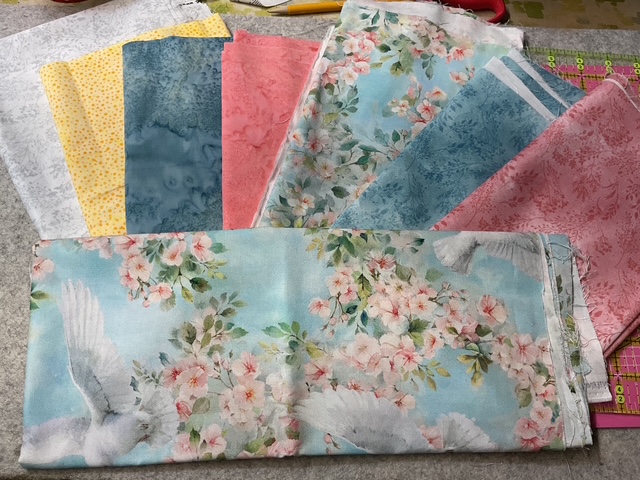 A collection of fabric pieces in various colors and patterns, including floral designs with birds and solid colors, spread out on a crafting surface.