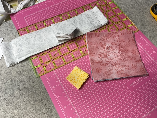 Various fabric pieces arranged on a pink cutting mat, including a long strip of white fabric, a square of reddish fabric, and a small yellow square with a polka dot pattern.