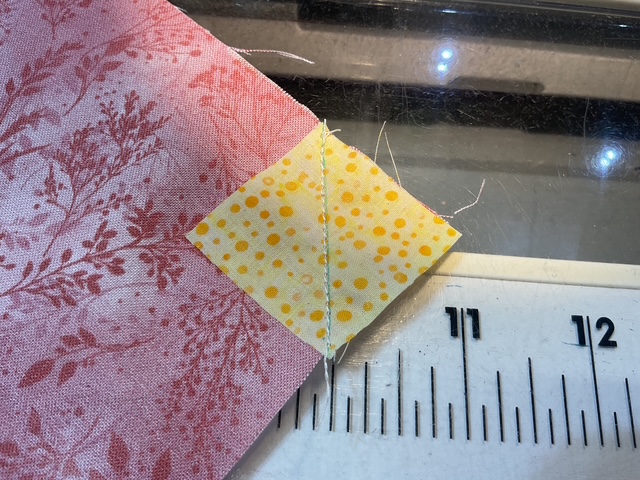 Close-up of two pieces of fabric being sewn together, one with a floral pattern in pink and the other a yellow fabric with polka dots. A ruler is visible in the background for measurement.