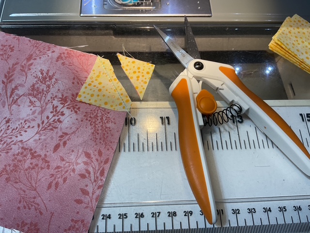A workspace featuring a pair of orange-handled scissors, two yellow fabric triangles, and a pink patterned fabric piece, all set on a cutting mat with a ruler.