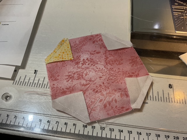 A partially sewn octagonal patchwork piece made of various fabrics, including pink and yellow, on a sewing table with measuring tools.