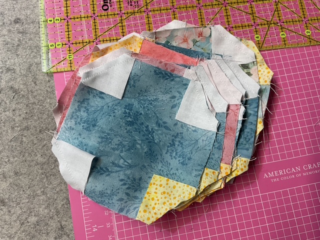 A stack of fabric pieces in various colors and patterns, including blue, pink, and yellow, arranged on a pink cutting mat with measuring grid.