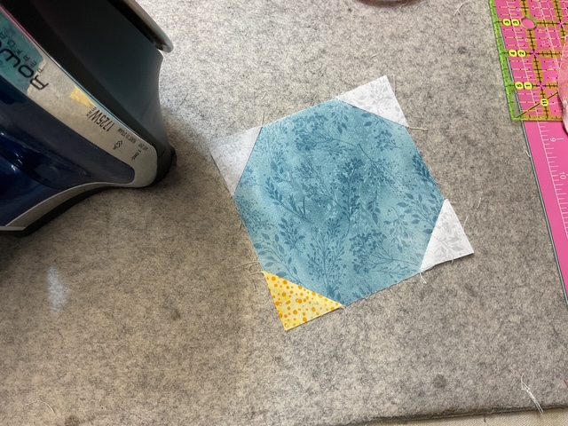 A sewing workspace featuring an iron next to a blue hexagonal quilt piece with floral patterns, set on a gray pressing mat.