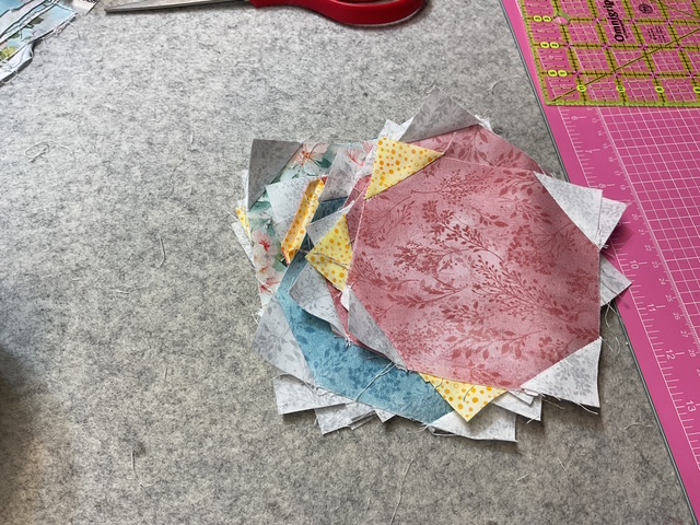 A stack of colorful quilt fabric pieces in various patterns, including shades of red, blue, yellow, and floral designs, on a gray cutting mat.