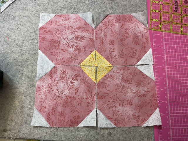 A quilt block featuring four patterned octagons in shades of pink with a central yellow diamond surrounded by white fabric, placed on a gray felt surface.