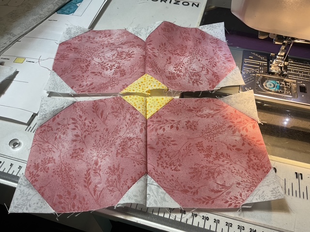 A close-up view of a sewing project showing four hexagonal fabric pieces arranged in a pattern, with pink floral fabric and a yellow center piece, placed on a sewing machine table.