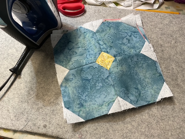 A blue and white hexagonal quilt block with a yellow center, placed on a gray fabric surface next to an iron and scissors.