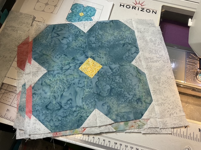 A sewing workspace featuring a pile of fabric pieces arranged in the shape of a flower, with blue and yellow patterns, set against a backdrop of quilting tools and a quilting machine.