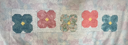 A fabric quilt with five large floral blocks arranged in a row, featuring colors of blue, pink, and a floral pattern, set against a pastel background.