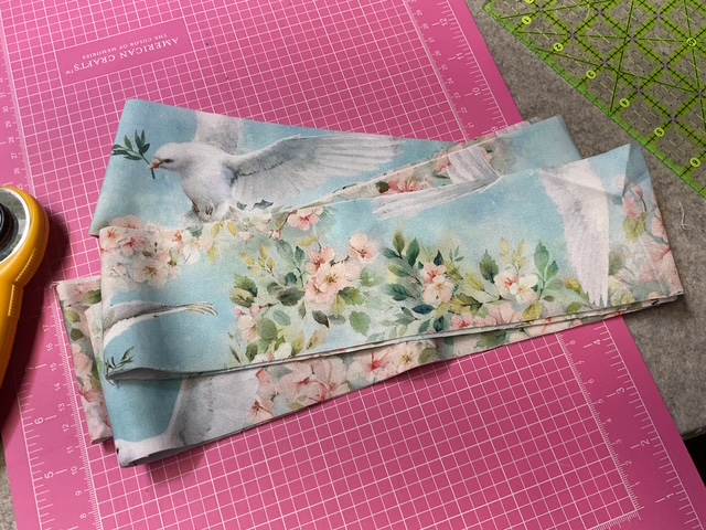 A stack of fabric strips featuring a pastel design of doves and floral patterns, placed on a pink cutting mat with a rotary cutter nearby.