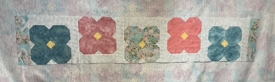 A quilt panel featuring a series of five floral patterns in varying colors including blue, pink, and green, with yellow accents in the center of each flower.