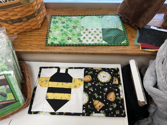 Two handmade quilted placemats displayed on a wooden surface. The top placemat features a green and plaid patchwork design with shamrocks, while the bottom placemat showcases a bee and beehive pattern in black, yellow, and white.