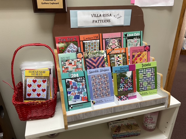 A display stand showcasing various quilting patterns from Villa Rosa, featuring designs such as 'Tea for 2', 'Lantern Hill', 'Love Cats', and more. A red basket with additional pattern booklets is also visible.