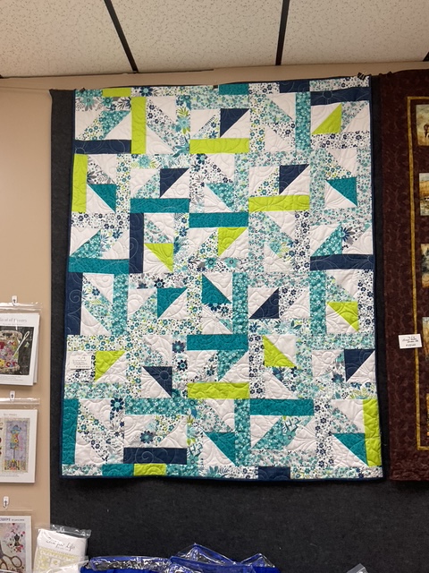A colorful quilt featuring a geometric pattern with shades of teal, navy, green, and white, displayed on a wall.