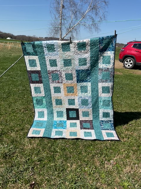 A colorful quilt hanging outside, displaying a pattern of squares in shades of blue, teal, and gray.