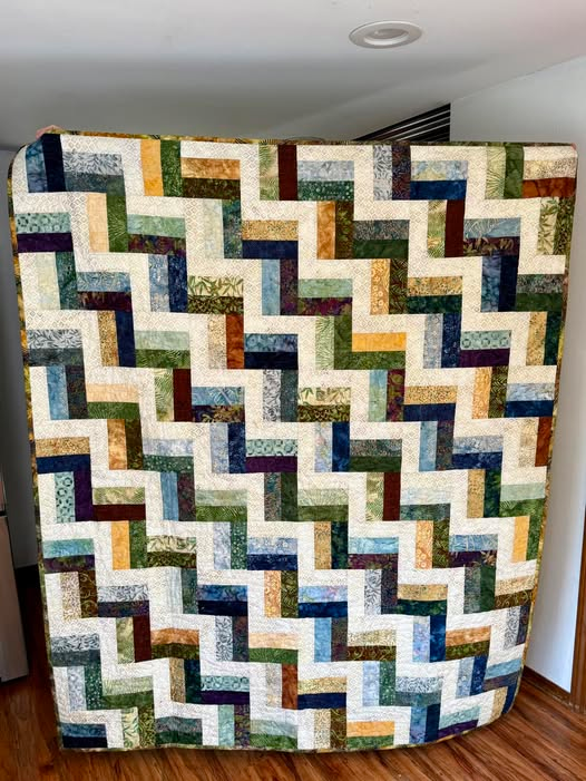 A colorful quilt displayed against a wall featuring a zigzag pattern with various shades of blue, green, yellow, and brown.
