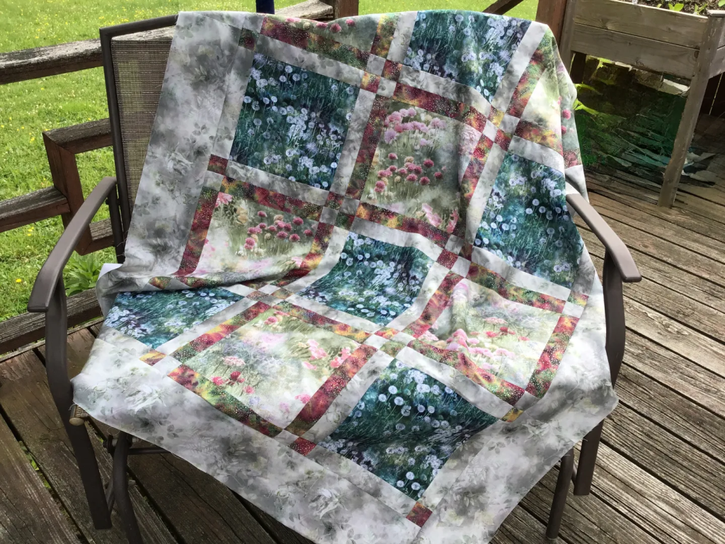 A colorful quilt draped over a chair, featuring floral patterns and a mix of green and pastel colors, resting on a wooden deck.