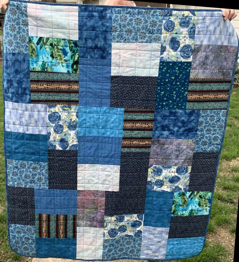 A colorful quilt featuring various shades of blue, green, and purple fabrics arranged in a patchwork design, with floral and geometric patterns.