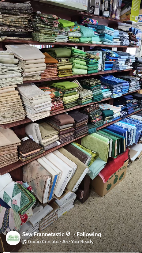 A display of neatly arranged fabric bolts on wooden shelves, showcasing a variety of colors and patterns, including greens, blues, and neutral tones, in a fabric store.