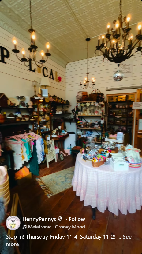 Interior of a quaint retail shop featuring vintage and decorative items. A round table with a pink checkered tablecloth is in the center, surrounded by shelves filled with various goods. Chandelier lights hang from the ceiling, and 'LOCAL' is displayed on the wall.