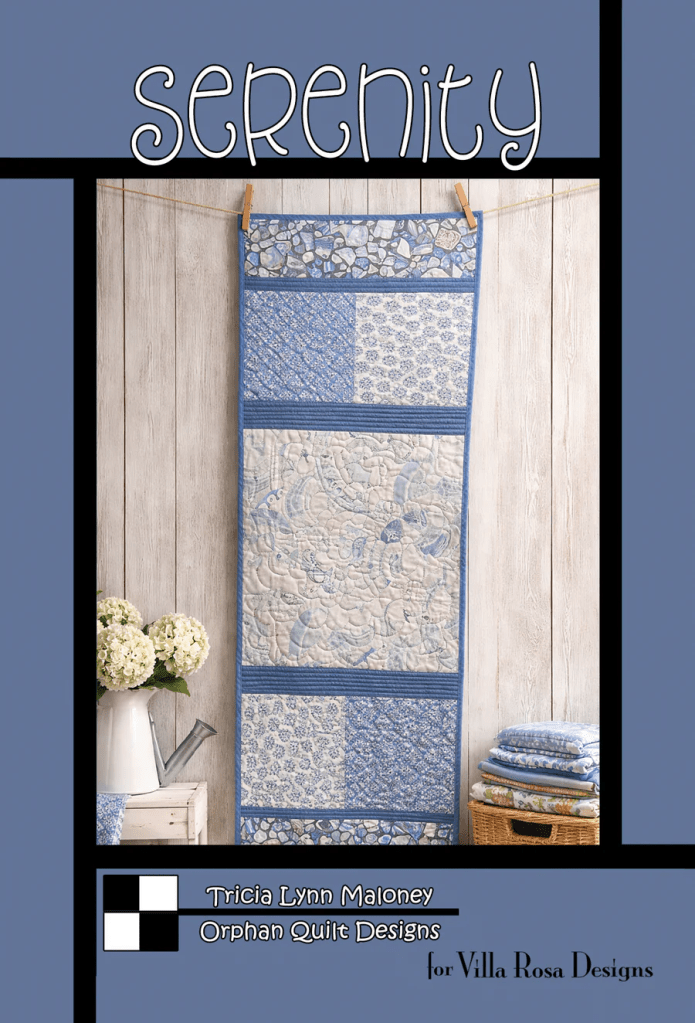 A wall-hanging quilt titled 'Serenity' featuring various blue and white fabric patterns, displayed against a wooden backdrop, accompanied by a vase of flowers and neatly folded quilts in a basket.