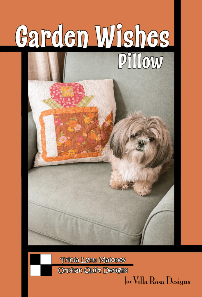 A small dog sitting on a couch next to a decorative pillow featuring a colorful flower pot design, with the text 'Garden Wishes Pillow' above it.
