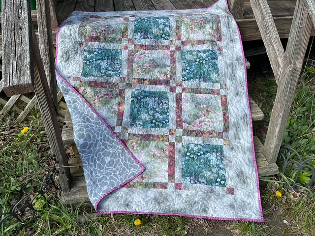 A colorful quilt with floral patterns draped over a rustic wooden bench. The quilt features squares of various floral designs and is bordered with a gray patterned fabric.