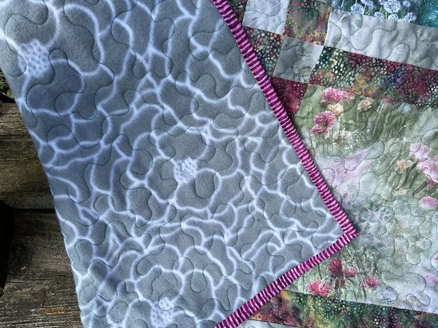 Close-up of a quilt featuring a textured gray fabric with a swirling pattern, edged with a pink and white striped binding, alongside a patchwork design of floral and textured fabrics.
