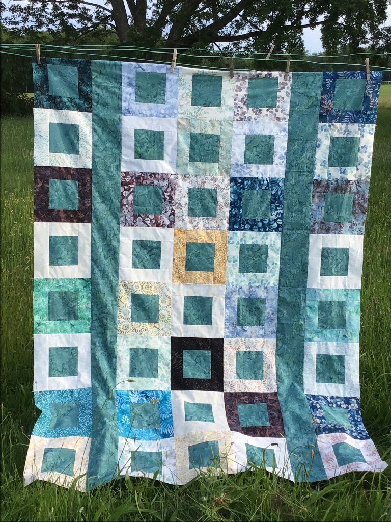 A colorful patchwork quilt hanging outdoors, displaying various shades of blue, green, and white fabrics arranged in a grid pattern.