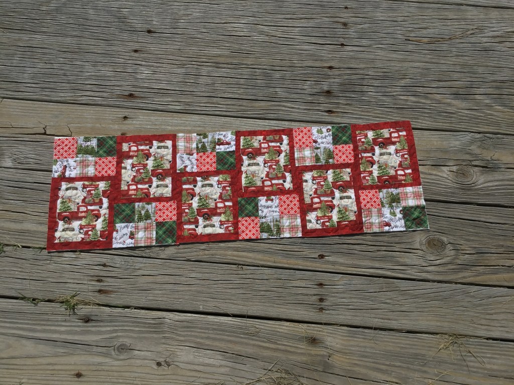 A patchwork table runner featuring red and green Christmas-themed fabrics, including vintage trucks, trees, and festive patterns, laid out on a wooden surface.