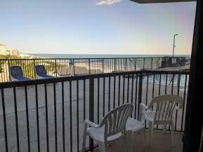 A balcony view overlooking the beach with two white chairs and a railing, featuring a clear sky and ocean in the background.