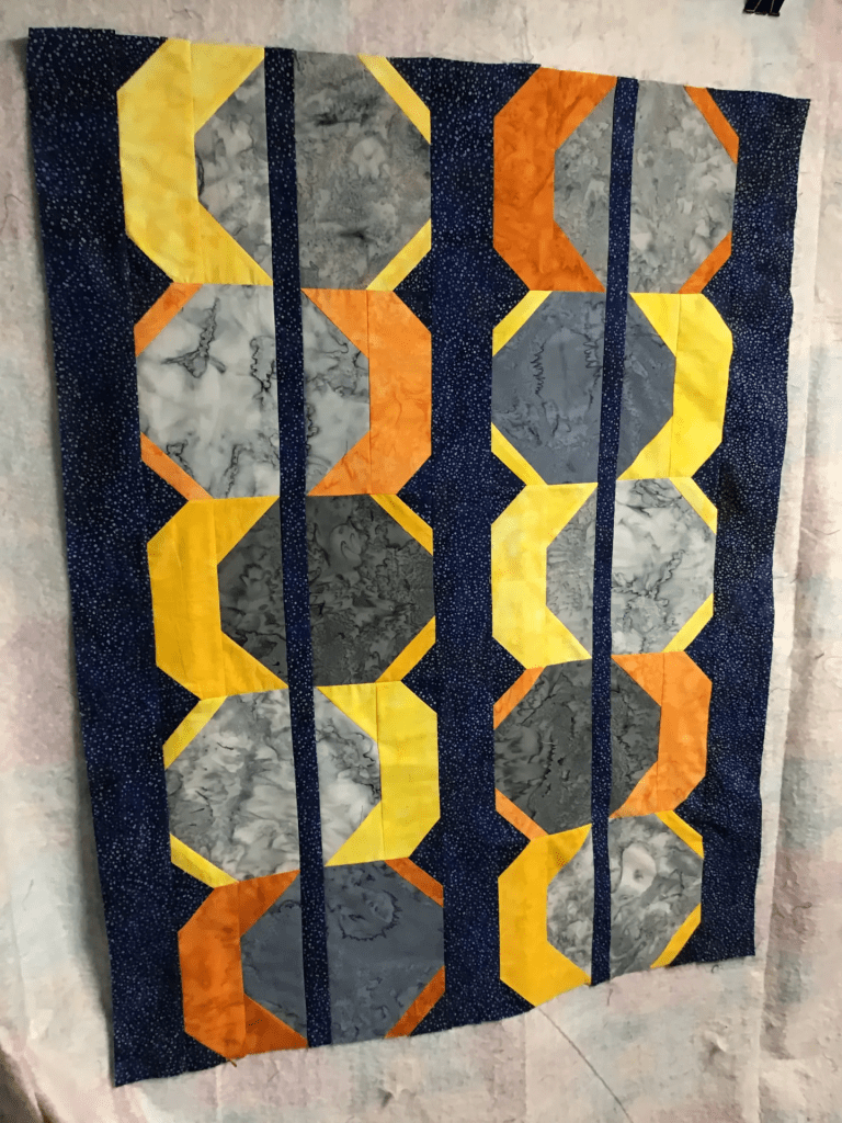 A colorful quilt featuring geometric patterns with yellow, orange, gray, and navy blue fabrics arranged in vertical strips.