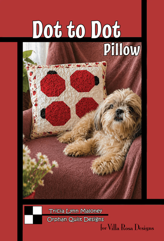 A decorative pillow featuring a red and black hexagonal design is displayed on a brown couch, accompanied by a small, fluffy dog resting beside it.